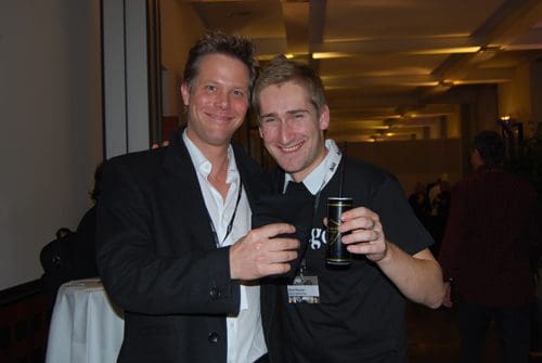 Two men standing together at an event, both smiling and holding drinks. One man is dressed in a black suit with a white shirt, while the other wears a black t-shirt. A social gathering is visible in the background.