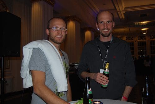 Two men standing at a table in a dimly lit event space. One man wears glasses and a light gray t-shirt with a sweater draped over his shoulder, while the other wears a black shirt and holds a beer bottle. Both appear to be enjoying the gathering.