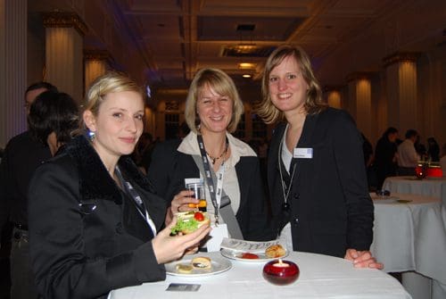 Three women standing at a table in a banquet setting, each smiling and dressed in formal attire. One woman holds a plate with a salad and appetizer. The background features elegant decor and attendees mingling.