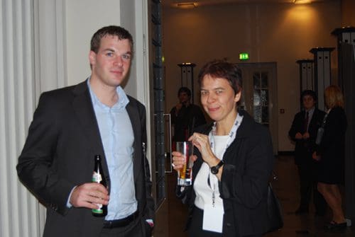 A man in a black suit and blue shirt stands smiling while holding a beer, next to a woman in a black blazer and white shirt, holding a glass with a drink. The background features a modern, elegant setting with dim lighting.