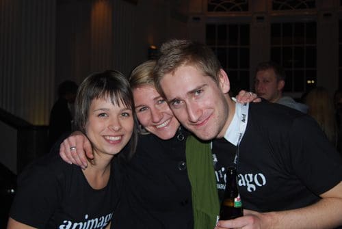 Three friends posing for a photo in a dimly lit venue. Two women and a man wearing matching black t-shirts with the word 'animago' are smiling, with the woman on the left resting her hand on the other's shoulder.