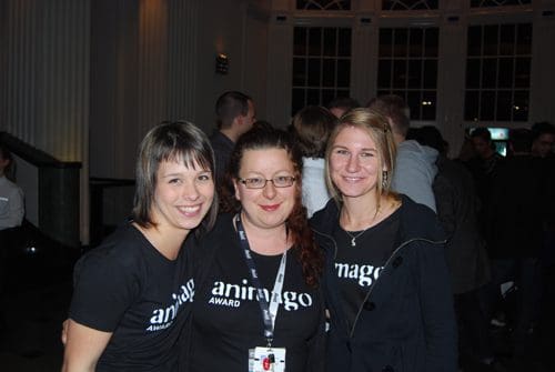 Three women smiling together at an event, all wearing black shirts with the text 'animago AWARD.' The background features a lively gathering of attendees in a well-lit space.