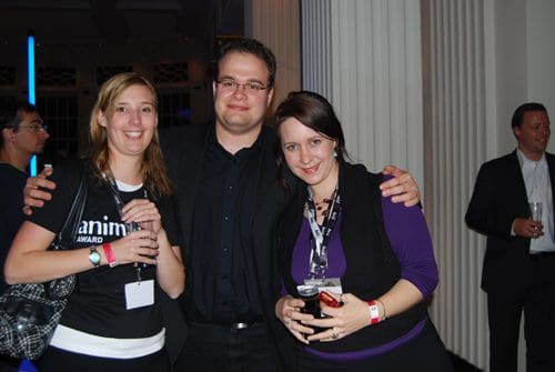 Three people posing for a photo at an event. The woman on the left, wearing a black shirt with "anim" text, holds a drink. The man in the center is wearing a black shirt and glasses, while the woman on the right is dressed in a black and purple outfit, also holding a drink. Soft blue lighting is visible in the background.