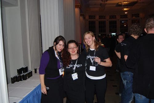 Three women posing together at an evening event. They wear black t-shirts with the word 'ninjago' on them. The background is filled with attendees, and a long table with drinks is visible to the left.