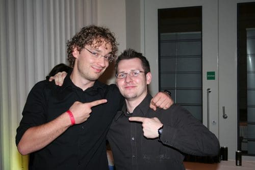 Two men wearing black shirts pose for a photo indoors, both smiling and pointing at each other. The man on the left has curly hair and glasses, while the man on the right has short hair and is clean-shaven. A soft light ambiance is visible in the background.