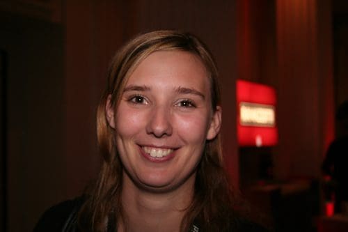 A smiling woman with long, light brown hair stands in a softly lit environment. A blurred red sign is visible in the background, suggesting a social or networking event.