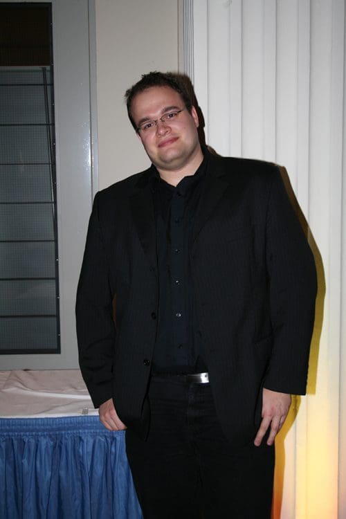 A man in a black suit and glasses standing confidently against a neutral-colored wall, with a soft light illuminating his side, creating a warm ambiance.