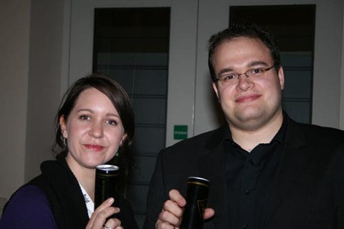 A woman and a man holding cans, smiling at the camera in a social setting. The woman wears a black vest over a purple shirt, and the man is in a black shirt with glasses, set against a backdrop of indoor lighting.