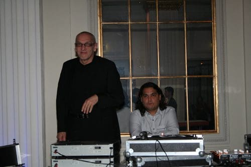 Two men are set up behind a DJ booth in a well-lit venue. One man, wearing glasses and a black outfit, stands beside a mixer, while the other, in a white shirt, sits focused at the controls. A large mirror reflects the atmosphere.