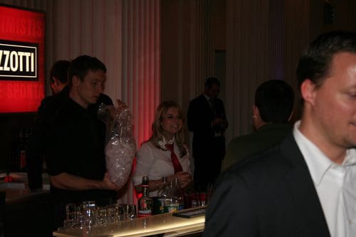 A lively bar scene with a female bartender smiling while holding a bag of ice, as a male colleague in a black shirt prepares behind the counter. Various glasses and bottles are arranged on the bar, illuminated by ambient lighting.