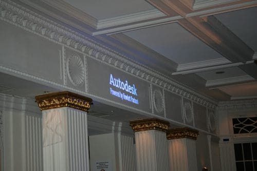 A dimly lit room showcasing a wall with decorative moldings, featuring a projection that reads "Autodesk" and "Powered by The Render Project". The scene includes ornate columns with gold accents.
