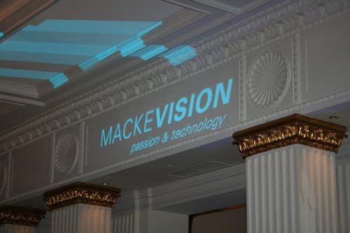 A projection of the word 'MackeVision' along with the tagline 'passion & technology' on an ornate wall with decorative gold accents and white columns in a well-lit interior space.