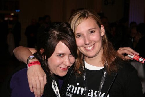 Two women smiling and posing closely together at an event, one wearing a black shirt with a logo and the other in a purple top. Both have lanyards around their necks, suggesting they are attendees or participants.
