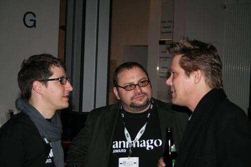 Three professionals in conversation at an event, wearing matching black t-shirts. The man on the left wears glasses and a scarf, while the man in the center has a beard and is dressed in a dark coat. The third man on the right talks animatedly, gesturing with his hand.
