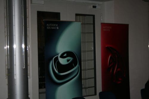 Two vertical banners displayed in a dimly lit room. One banner, in teal, features the Autodesk 3ds Max logo, while the other, in red, showcases the Autodesk Maya logo. The background includes a glass door and metal columns.