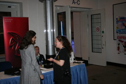 Two women engaged in conversation at a conference or event entrance, with a promotional banner for Autodesk Maya B in the background. A seated woman is visible at a table nearby, and doors labeled A-C are open.
