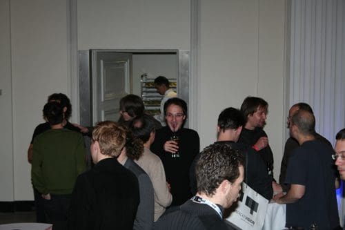 A group of people mingling at a social gathering, with some individuals chatting and smiling. One person in the foreground holds a drink, while others are engaged in conversation. In the background, a door leads to another room with shelves visible.