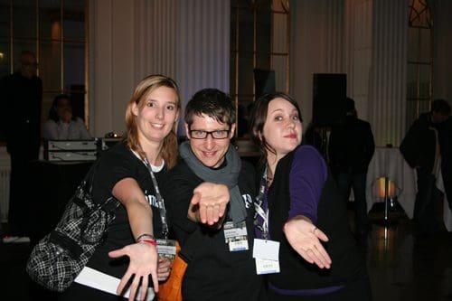 Three friends posing together at a social event, smiling and extending their hands forward in a playful manner. They are dressed casually and are in a well-lit indoor space with a festive atmosphere.