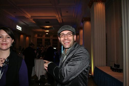 A man in a gray suit and hat smiles while pointing to the right, standing next to a woman in a dark outfit at a dimly lit reception. The background features elegant decor and guests mingling.