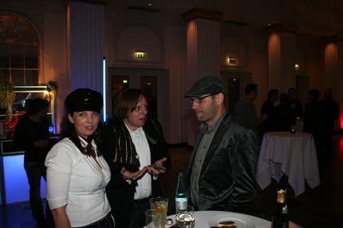 Three individuals engaged in conversation at a social event in a well-lit venue. The woman on the left wearing a black hat smiles warmly, while the man in the center gestures expressively. The man on the right, wearing glasses and a cap, listens attentively.