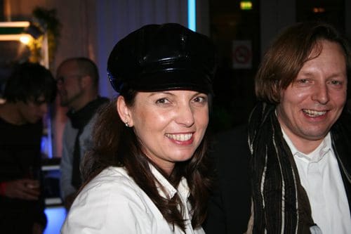 A woman with a black cap and a white shirt smiles at the camera, while a man with long hair wearing a black coat and a white scarf stands beside her, both in a lively event atmosphere with soft blue lighting.