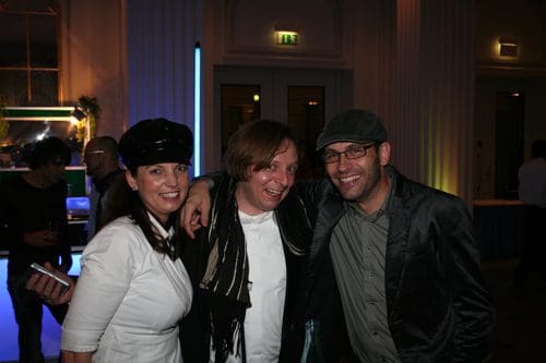 Three people smiling and posing for a photograph at a social event. The woman on the left is wearing a black hat and a white shirt, while the man in the middle has long hair and a black scarf. The man on the right is wearing glasses and a gray jacket.