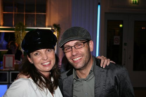 A woman wearing a black hat and a white blouse smiles next to a man in a gray hat and glasses, both posing happily in a brightly lit venue with colorful decor.