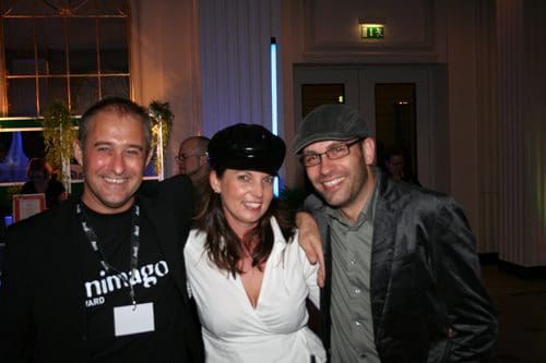 Three people posing together at an event, with one man wearing a black t-shirt, another in a gray suit, and a woman in a white blouse and black hat, all smiling against a backdrop of soft, ambient lighting.