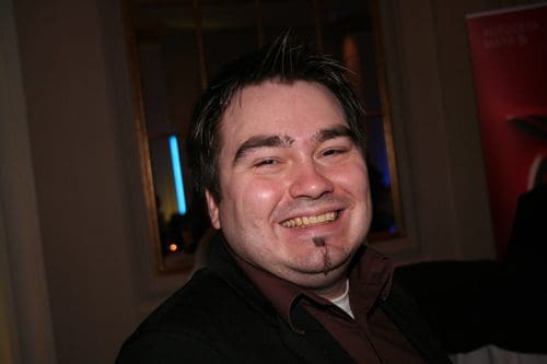A man with short, dark hair and a light beard smiling brightly at the camera. He is wearing a dark jacket over a collared shirt, standing in a room with soft lighting and decorative elements in the background.