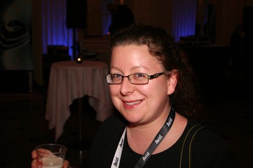 A woman with glasses smiling while holding a plastic cup at a social event. She has curly hair and wears a black jacket with a white lanyard. Soft, ambient lighting and a blurred background suggest a festive atmosphere.