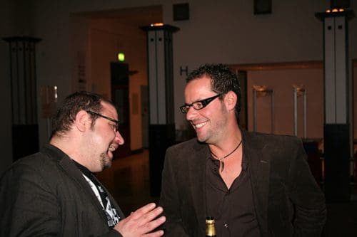 Two men engaging in a lively conversation at a social gathering. Both are wearing glasses and casual attire, with a relaxed atmosphere highlighted by soft lighting in the background.