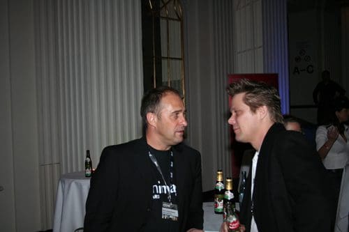 Two men in black blazers engaged in conversation at a social event, with bottles on a table nearby. They appear to be discussing something intently, set against a backdrop of elegant decor.