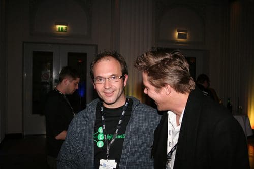 Two men standing close together in a lively indoor event. One man, wearing glasses and a black shirt with a green logo, smiles at the camera, while the other, in a black coat, laughs and leans towards him. A blurred figure can be seen in the background.