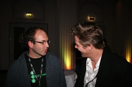 Two men in conversation at a social event, one wearing glasses and a dark t-shirt with a graphic, the other in a white dress shirt and black blazer, both smiling, with softly lit background decorations.
