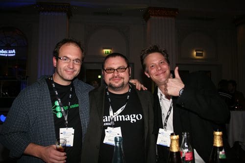 Three men posing together at an evening event. They are dressed in casual and semi-formal attire, smiling and appearing relaxed. Bottles are visible on the table in front of them, adding to the festive atmosphere of the gathering.