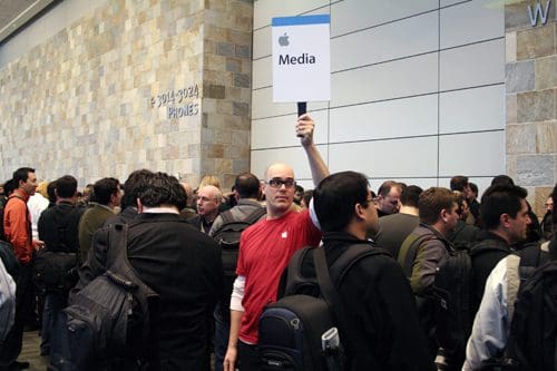 A crowd of people gathered in a large venue, with a man in a red shirt holding a sign that reads 'Media' with an Apple logo. The atmosphere is busy, with attendees carrying bags and engaged in conversation.