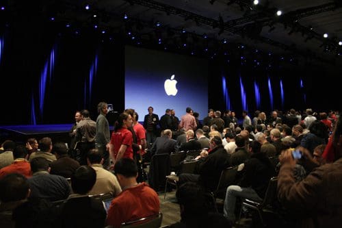 A crowded auditorium at a tech event with attendees gathering and conversing, while a large screen displays the Apple logo. The stage is illuminated with blue lighting, setting a dynamic atmosphere.