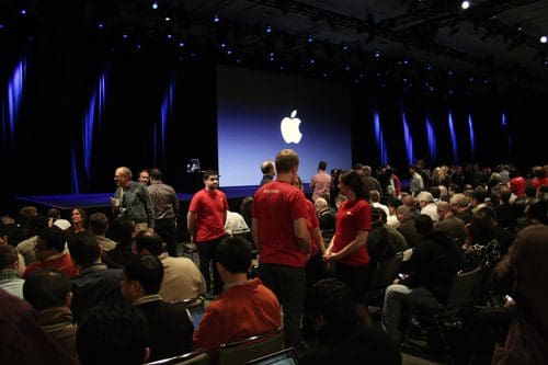 An auditorium filled with an audience awaiting a presentation, featuring attendees in casual attire and staff in red shirts. The stage is set with a large Apple logo displayed on a screen, illuminated by bright blue lights.