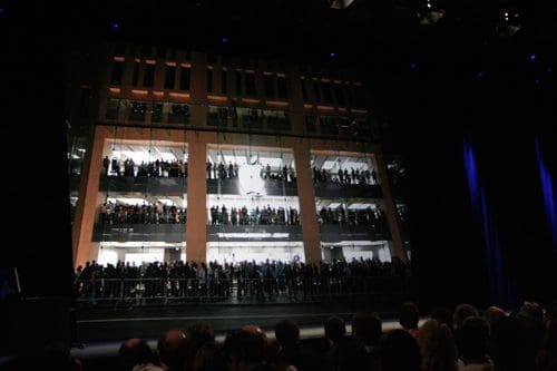 A large crowd of people gathering on multiple levels of a modern building, with some individuals waving. The interior is brightly lit, contrasting with the darkly lit auditorium in the foreground.