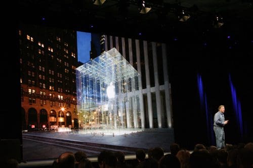 A large screen displays a modern glass Apple Store at night, illuminated and reflecting city lights. In the foreground, a speaker stands on stage with a captivated audience seated below, showcasing urban technology innovation.