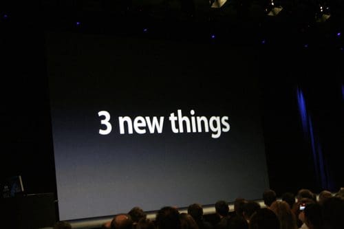 A large screen displaying the text '3 new things' during a presentation, with an audience seated in the foreground, capturing a moment of anticipation in a dimly lit auditorium.