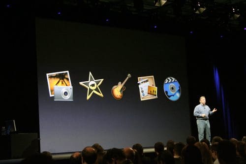 A speaker presenting on stage in a darkened auditorium, with large colorful icons representing creative software on a screen behind him. An audience is visible, attentively watching the presentation.