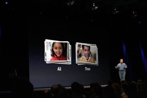 A speaker standing on stage presenting two children's photos, one labeled 'Ali' and the other 'Theo.' The backdrop is dark, and the audience is partially visible in the foreground.