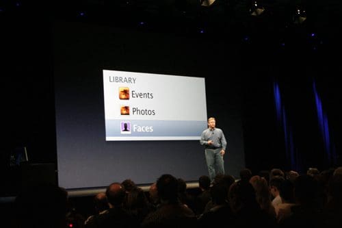 A speaker presenting on stage in front of a large screen displaying a computer interface with options for 'Library,' 'Events,' 'Photos,' and 'Faces,' while an attentive audience observes.