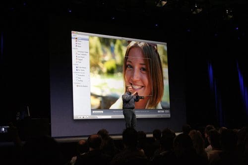 A speaker on stage holding a camera, with a large display behind showing a smiling woman outdoors. The audience is seated, focusing on the presentation in a well-lit auditorium.