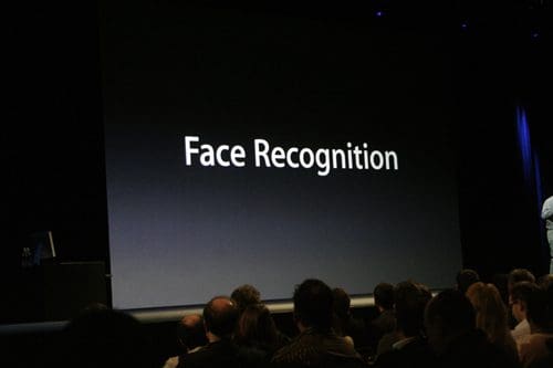A large screen displaying the text "Face Recognition" in bold white letters, while an audience of people is seated in the foreground, facing the stage.