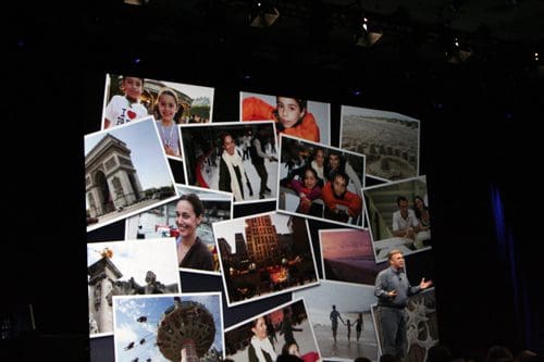 A speaker stands on stage in front of a large backdrop featuring a collage of various travel photos, including iconic landmarks and smiling individuals, creating a vibrant presentation atmosphere.