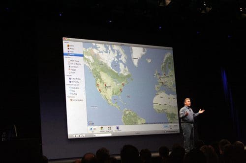 A speaker gesturing towards a large presentation screen displaying a map of North America. The audience is seated in a dimly lit theater with a focus on the speaker and the map.