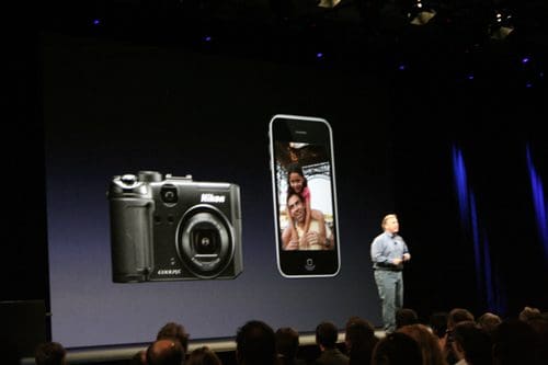 A speaker presenting on stage at a conference, with a large screen displaying a Nikon camera and a smartphone showing a photo of two people. The audience is visible in the foreground, engaged in the presentation.