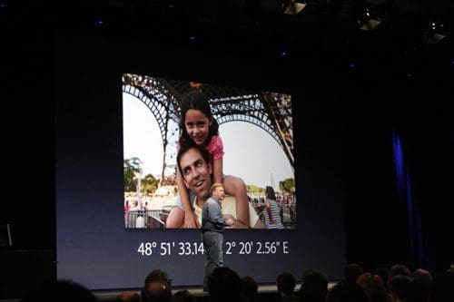 A man smiling while holding a girl on his shoulders, both near the Eiffel Tower, displayed on a large screen during a presentation. The audience is visible seated in the darkened space.